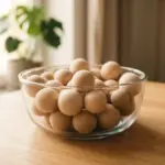 A glass bowl filled with wooden spheres sits on a wooden table near a plant by a window