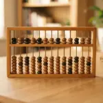 A wooden abacus rests on a light-colored wooden desk in a blurred room with a plant and books